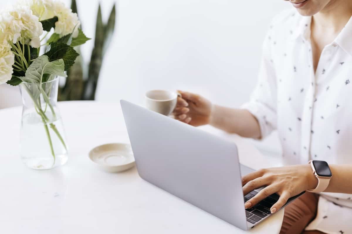 Woman in a white shirt sitting in front of a laptop looking at a homepage. There are flowers on the table and she has coffee in her hand.
