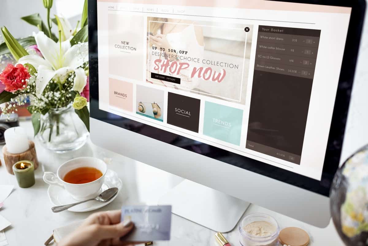 Computer screen with online shopping and the large words 'shop now' surrounded by flowers and tea on a desk and a hand holding a credit card.