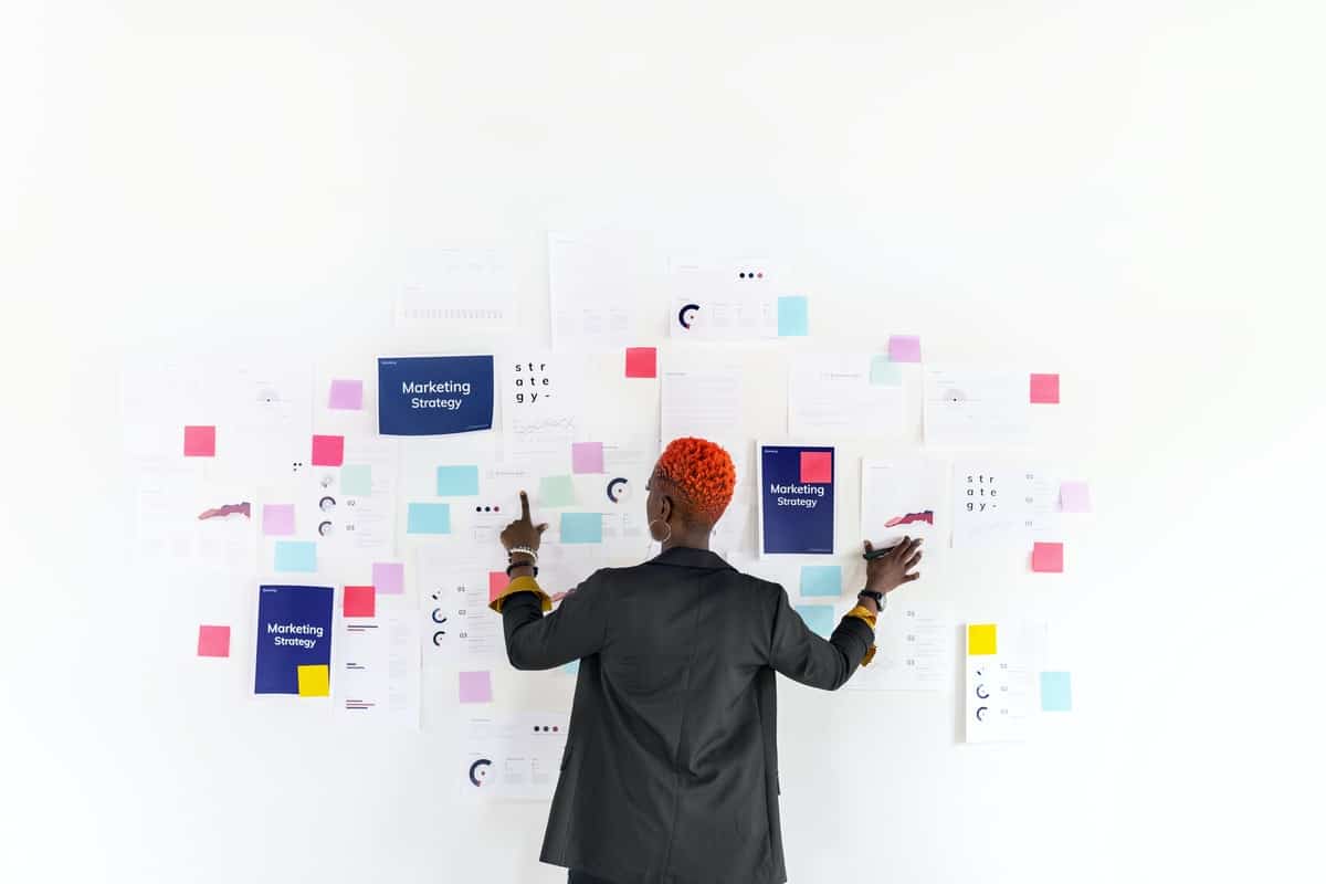 African american woman with a black blazer and short red hair looks at a white wall of sticky notes representing marketing strategy.