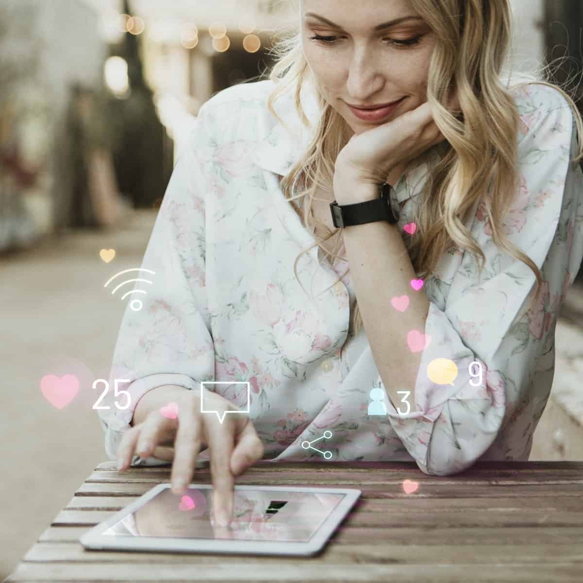Blond woman sits at a wooden table outside and uses her tablet. She has one hand under her chin and the other hand on the tablet screen.