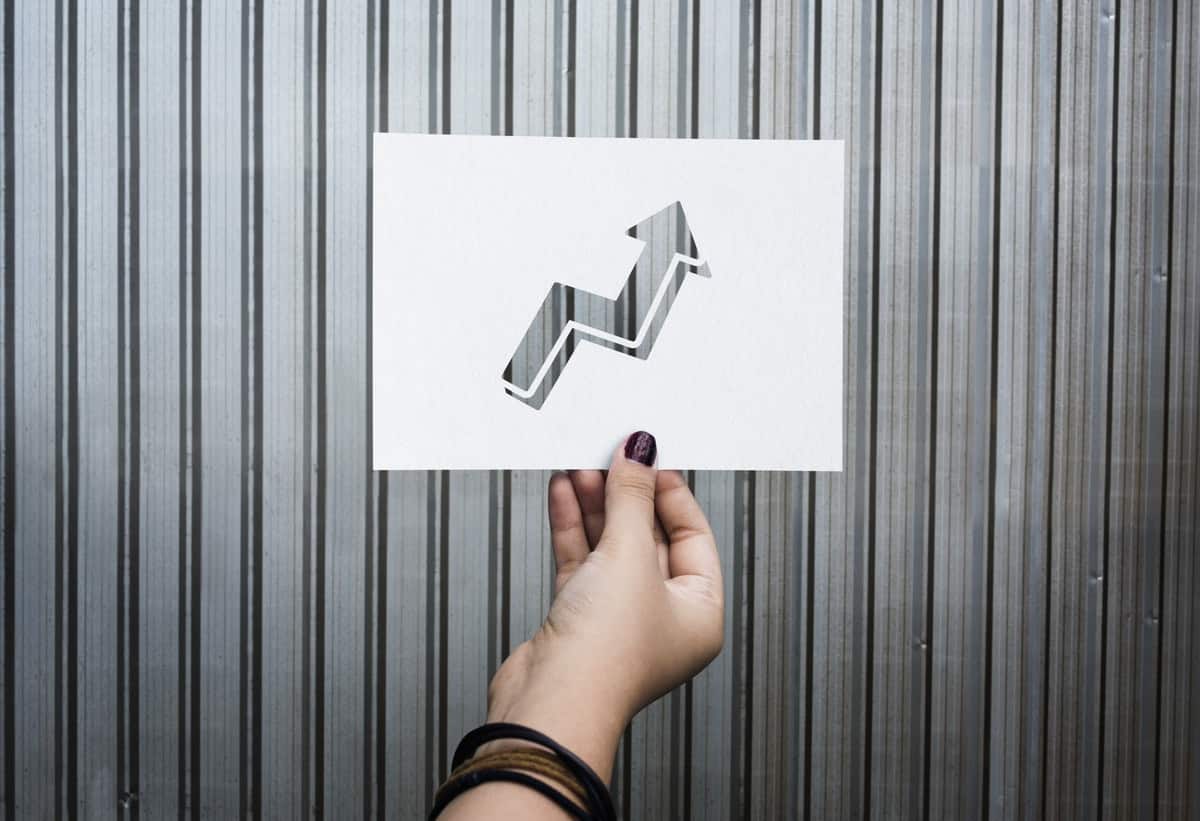 Woman's hand holds a rectangle piece of paper against a grey wall. The paper is white and has an upward pointing arrow cut out of it indicating growth-driven design..