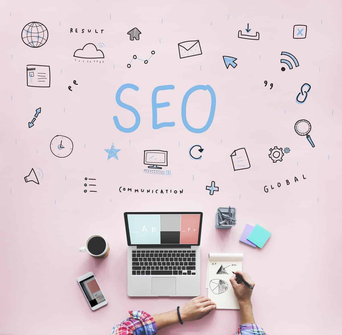 The word SEO in large letters with digital icon symbols on a pink background over a laptop, cup of coffee, and office supplies and two hands writing on a notepad.